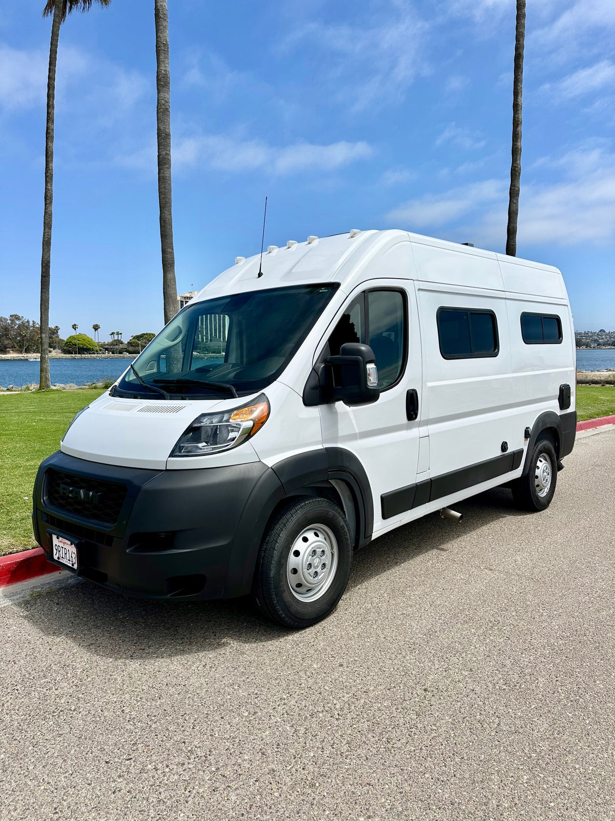 White Ram ProMaster camper van, an RV rental, parked by a sunny California waterfront with palm trees.