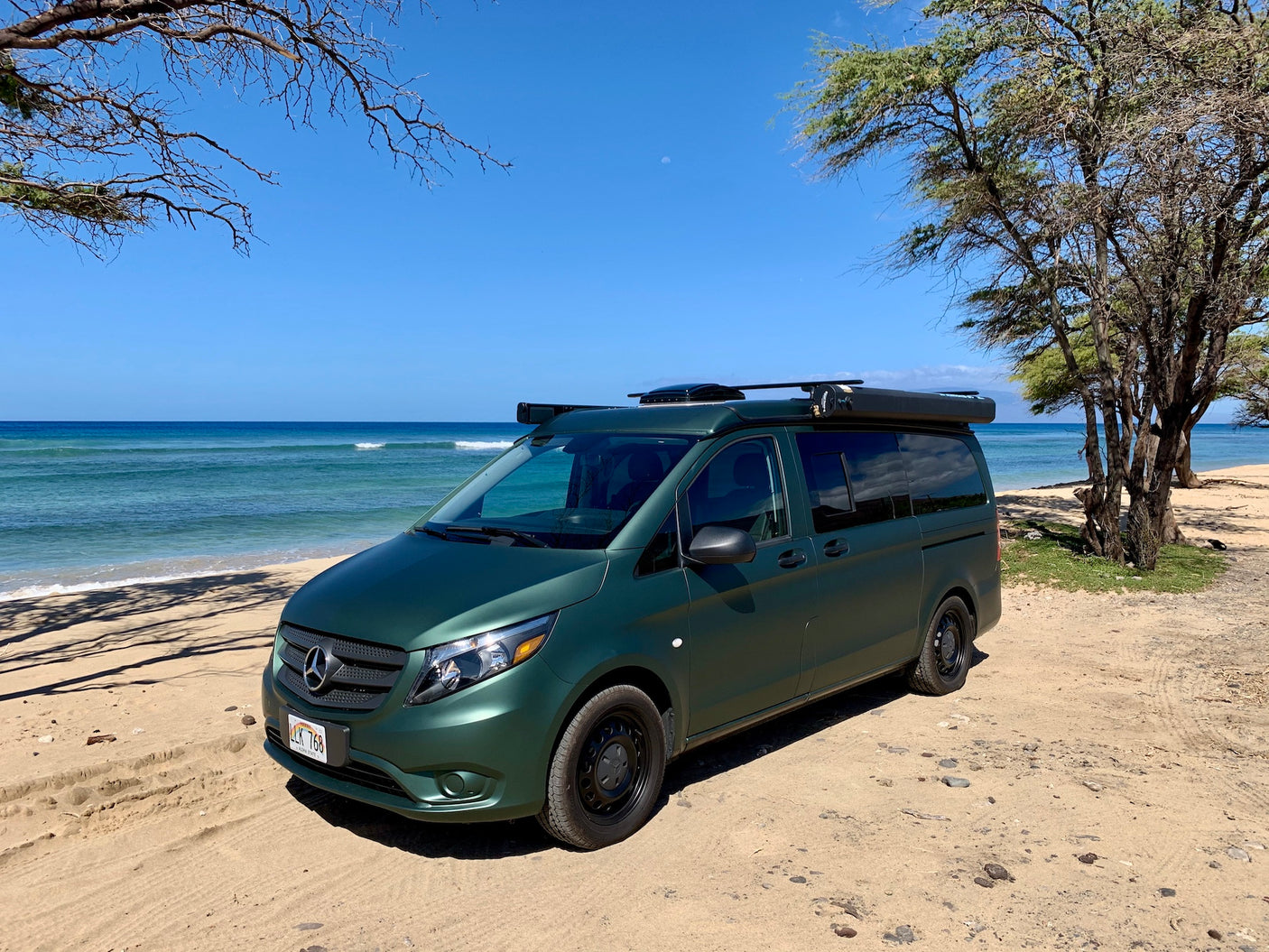 This green Mercedes Metris camper van, an ideal rv rental, is parked on a sandy Hawaiian beach by the ocean.