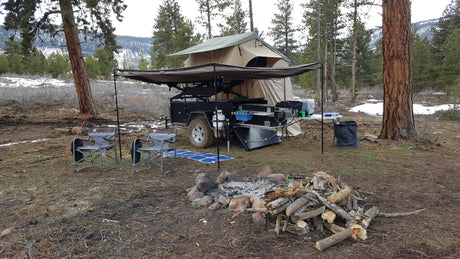 Adventure-ready RV rental: an overland camper trailer with rooftop tent, awning, pull-out kitchen, and fire pit in a snowy forest camp.