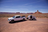 White pickup truck with bikes and a teardrop RV rental camper parked in a vast red rock desert under a blue sky, ready for adventure.