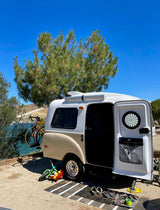 Lakeside rv rental teardrop camper with an open door, bike rack, and outdoor gear, perfect for a sunny camping and travel experience.