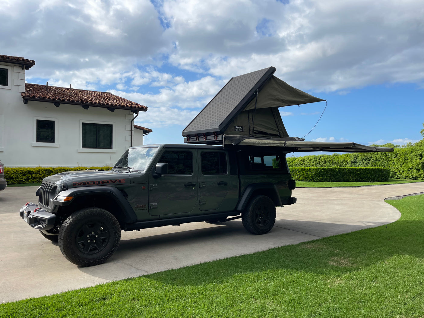 A dark green Jeep Gladiator, equipped with an open rooftop tent and side awning, parked on a driveway. Ideal for an RV rental adventure.