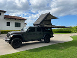 A dark green Jeep Gladiator, equipped with an open rooftop tent and side awning, parked on a driveway. Ideal for an RV rental adventure.