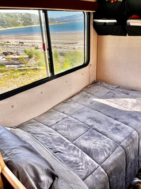 Cozy RV rental interior with a gray made bed and a window view of a tranquil beach and ocean, perfect for a getaway.