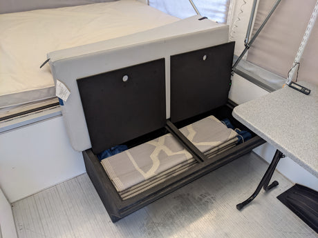 RV rental interior showcasing smart storage: A bed, an open bench with folded mats, and a side table maximize space.