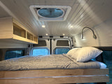 Camper van interior of an RV rental: a built-in bed, wood cabinets, ceiling fan, and reading lights create a cozy space.