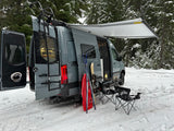Snowy RV rental setup: A camper van with an awning, ladder, skis, sled, chairs, and table, ready for winter adventure in a forest.
