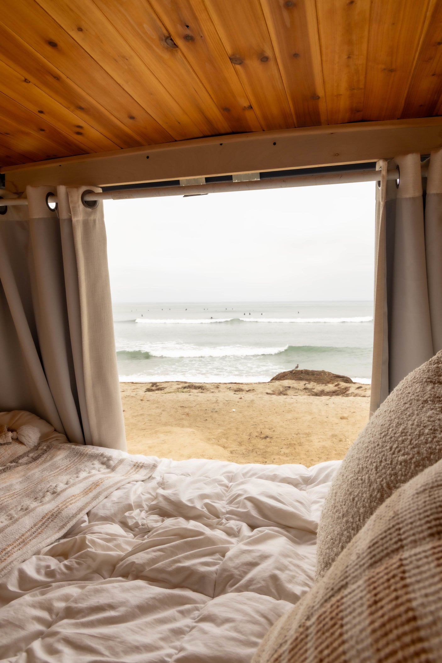 Cozy RV rental bed with white linens, overlooking a beach with waves, surfers, and a distant seagull.