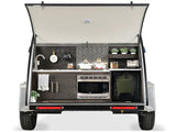 RV rental camper trailer's rear hatch reveals a compact outdoor kitchen: sink, stovetop, microwave, storage.
