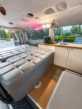 Cozy RV rental interior features a large gray bed, compact kitchenette with sink, and a bright 'inspire' neon sign.