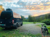 Dark RV rental van parked on gravel. Two large dogs watch a vivid sunset over hills and trees, perfect for outdoor adventure.