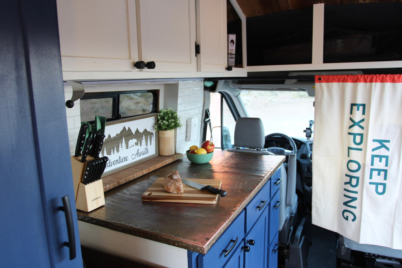 A compact RV rental kitchen boasts white and blue cabinets, a unique metal countertop, and 'Adventure Awaits' decor. — photo 9