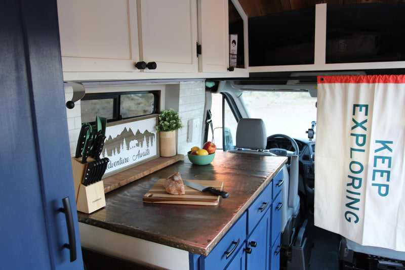 A compact RV rental kitchen boasts white and blue cabinets, a unique metal countertop, and 'Adventure Awaits' decor. — photo 9