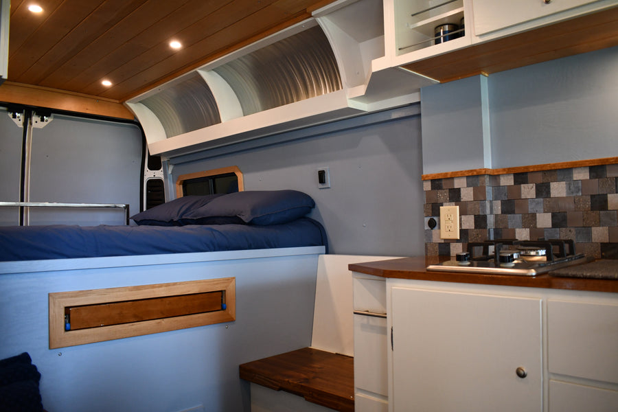 Experience comfort in this RV rental! Features a cozy bed, compact kitchen with a tiled backsplash, and smart overhead storage.