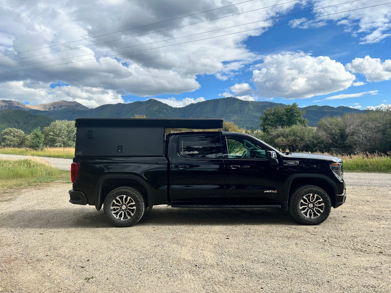 Black GMC Sierra AT4 truck with adventure truck camper on gravel, mountains in background. Ideal RV rental for overlanding. — photo 3