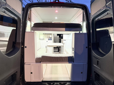 View of an RV rental camper van's interior from the open rear doors, revealing a cozy bed, dining area, and compact living space.