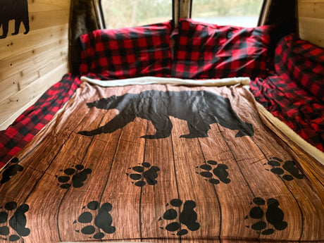 Rustic RV rental bed featuring a bear-themed blanket on wood-look fabric & red plaid pillows, ideal for nature getaways.