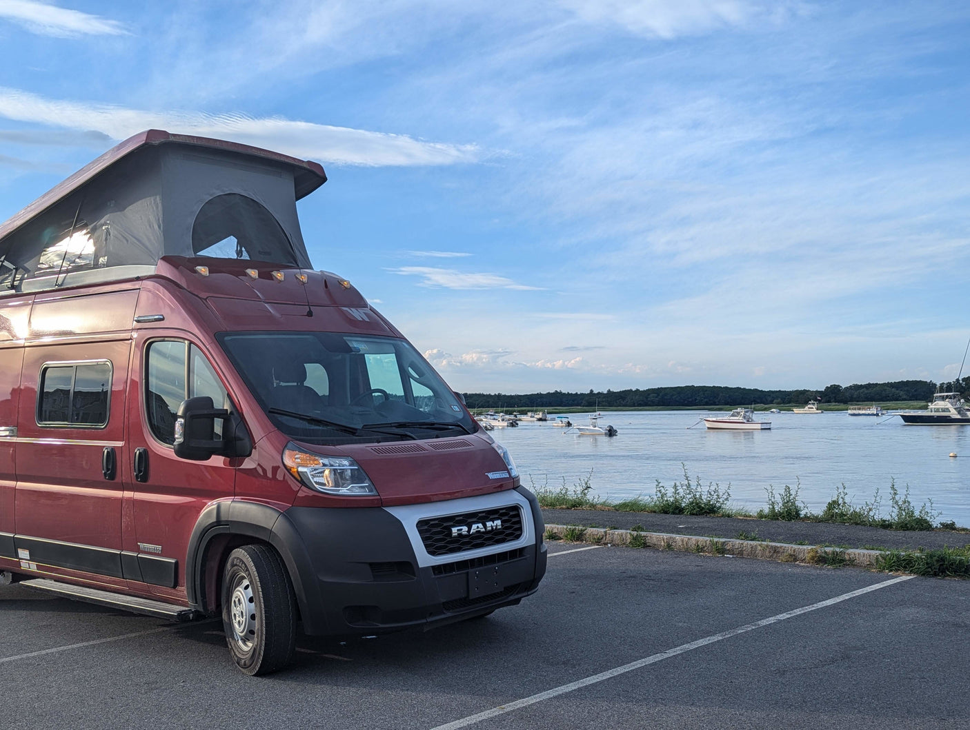 A red pop-top camper van, an RV rental, is parked on a lot beside a bay filled with boats. Perfect for scenic travel!