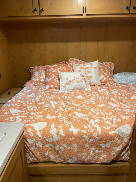 RV rental interior: cozy bedroom featuring a peach-patterned quilt, decorative pillows, and overhead wooden cabinets.