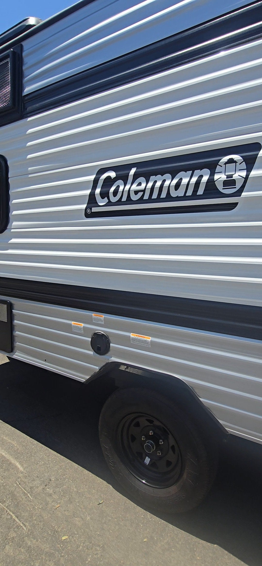Side of a new Coleman RV, showcasing its white ribbed exterior, logo, and tire. Ideal for an rv rental.