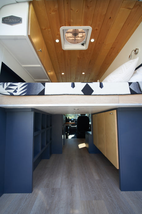 Experience this stylish RV rental! Elevated bed, wood ceiling, ample dark blue storage, and light flooring create a cozy mobile home.
