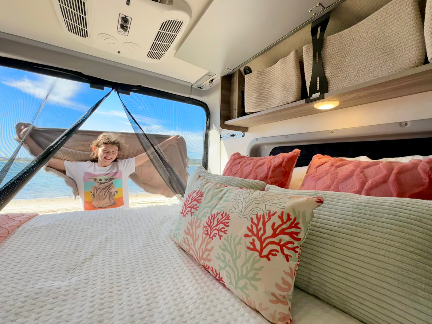 Enjoy stunning beach views from your comfortable RV rental. A happy girl smiles from inside, surrounded by cozy pillows.