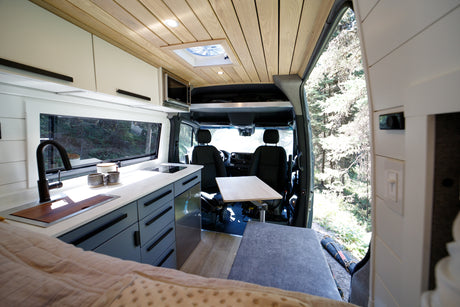 Spacious rv rental camper van interior with a modern kitchen, comfortable bed, and front table, all boasting forest views.