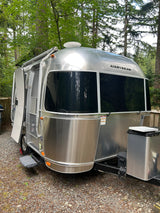 This gleaming Airstream travel trailer, ideal for an RV rental, is parked on gravel among tall trees, with its door open.