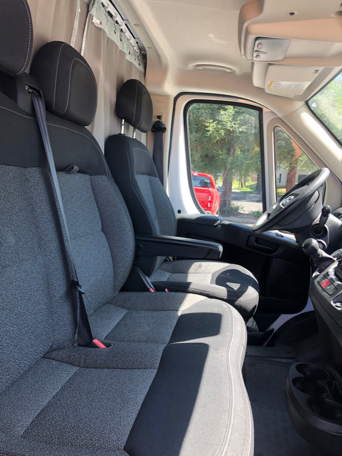 RV rental van's front cabin: gray and black seats, steering wheel, ceiling track for storage/curtains. A red truck is outside.