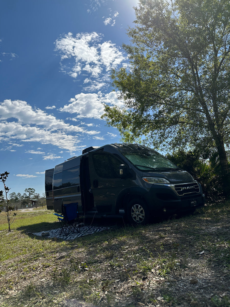 Peaceful camping scene with an RV rental. Dark gray camper van parked under a tree on a sunny day.
