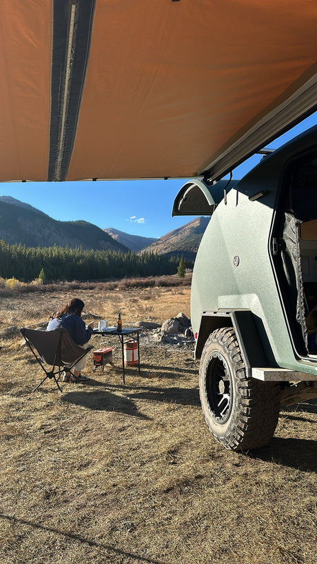 Camping with an RV rental: Person enjoys a meal beside a green teardrop trailer and awning in a mountain valley.