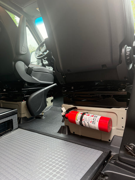RV rental van interior showing front seats, grey textured flooring, and a red fire extinguisher securely mounted by the passenger seat base for safety.