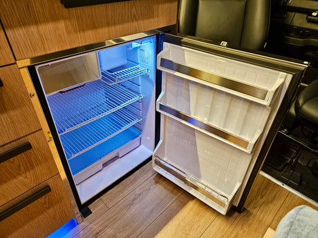 Open compact fridge in an RV rental, featuring blue interior light, wire shelves, and door storage within wood cabinetry.