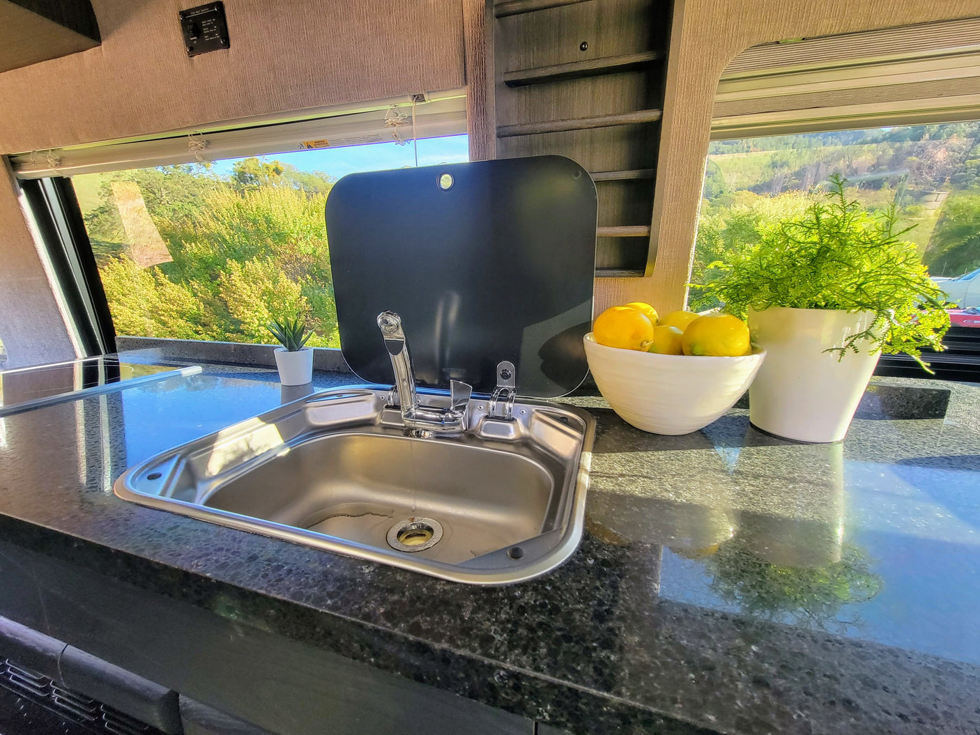 Sunny RV rental galley: modern sink, dark countertop, and scenic window view of vibrant green nature.