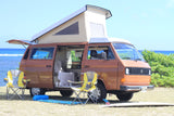 Brown VW pop-top camper van rv rental parked by the Hawaiian ocean with chairs & surfboard, ready for adventure.