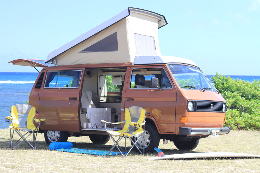 Brown VW pop-top camper van rv rental parked by the Hawaiian ocean with chairs & surfboard, ready for adventure.