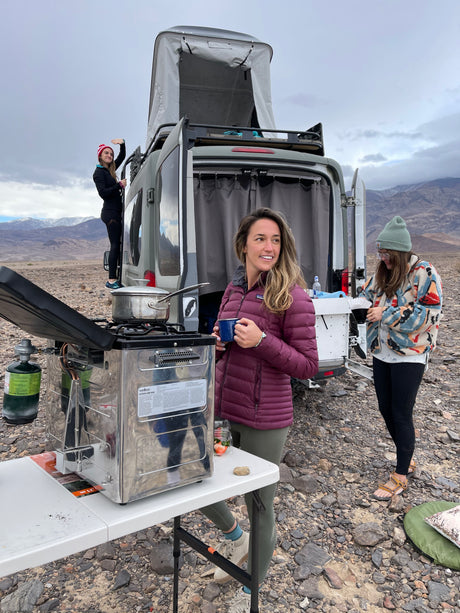 RV rental trip! Friends enjoy outdoor cooking & coffee by their camper van. One waves from the ladder, mountains behind.