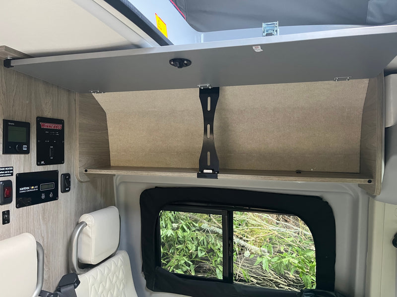 View inside an RV rental: open overhead storage, Winnebago & Truma control panels, and a window showing foliage. — photo 17