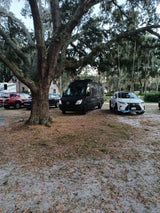 Black Sprinter van & white RV rental parked under a large tree, alongside other vehicles on a dirt lot.