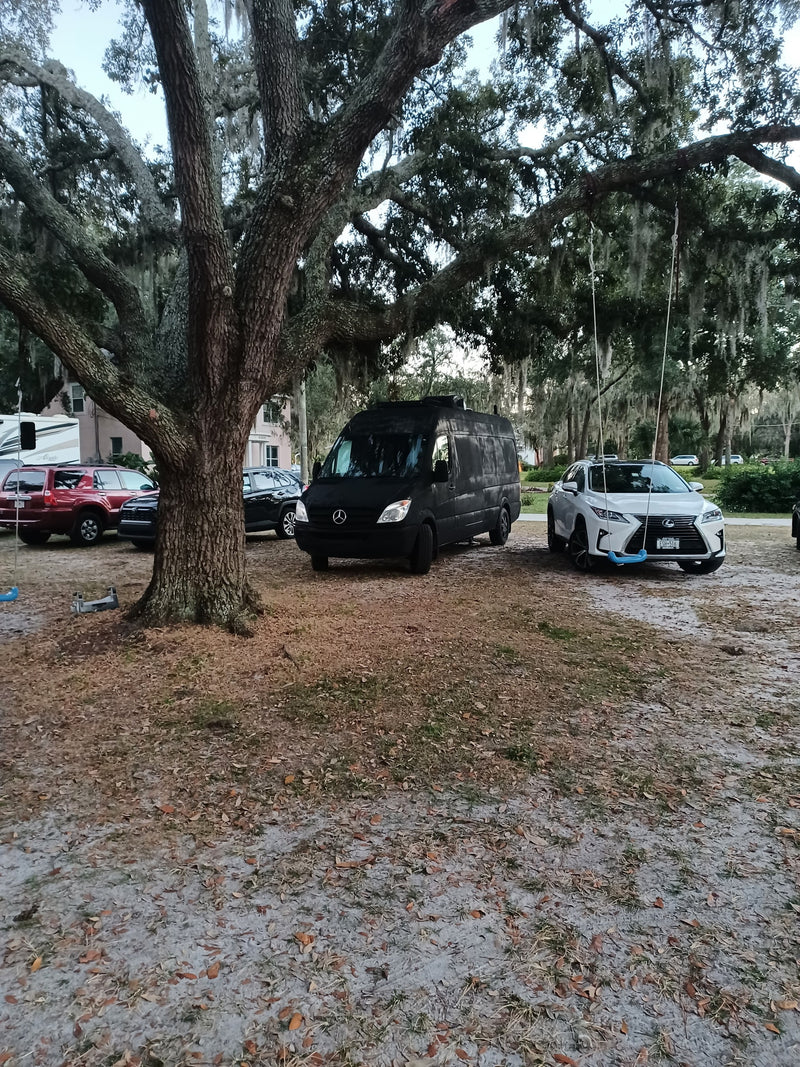Black Sprinter van & white RV rental parked under a large tree, alongside other vehicles on a dirt lot. — photo 6