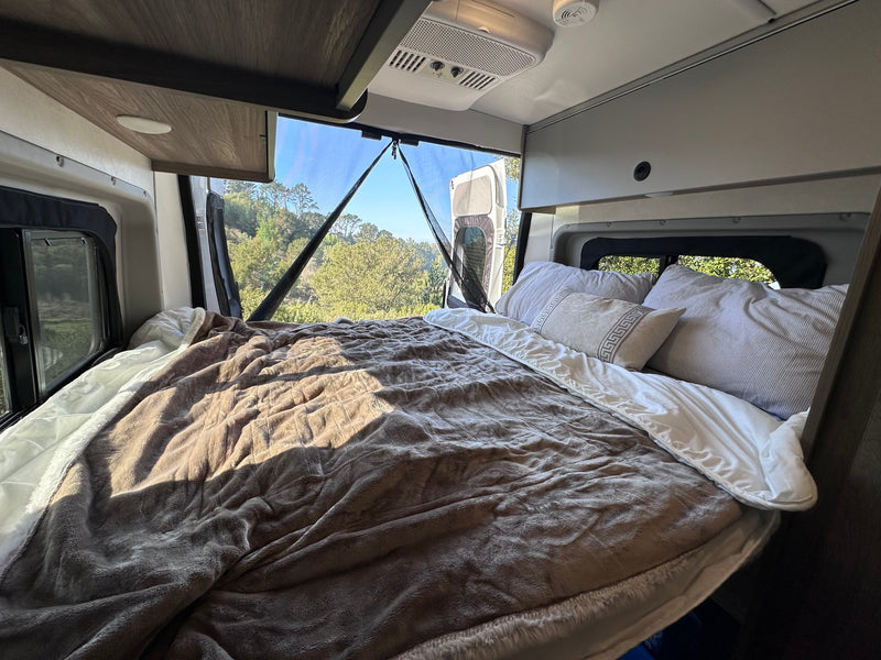  waves crash on rocks.|Peering through an RV rental's screen door at a serene natural landscape with lush green hills and trees. Van camping bliss!|RV rental interior: A cozy bed with a brown blanket and pillows offers serene views of green trees through the back windows.|Compact RV rental kitchen with a stainless steel sink — photo 5