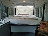 Cozy RV rental interior with a bed, small sink, and open back doors revealing a dry, grassy hill.