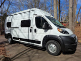 Bright white Winnebago Solis rv rental van parked on paved ground, surrounded by tall, bare trees under a clear blue sky. Ready for adventure.