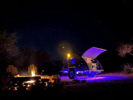Cozy night camping: a glowing teardrop RV rental, two chairs by a crackling fire under a clear dark sky.