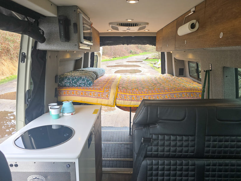 This RV rental interior shows a bed extending through open back doors to a muddy road. A kitchen counter and passenger seat are visible. — photo 2