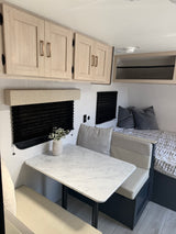 Light and airy rv rental interior featuring a dinette with a marble-look table, overhead cabinets, and a cozy bed with pillows.