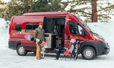 RV rental winter trip: A couple with snowboard & skis relaxes by their red camper van in a snowy forest scene.