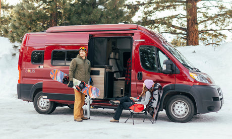 RV rental winter trip: A couple with snowboard & skis relaxes by their red camper van in a snowy forest scene.
