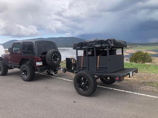 This camping trailer with a red rooftop tent & tarp is an excellent RV rental option for forest adventures.|This black off-road camping trailer with a tent frame is parked in a grassy forest. Great for your next RV rental adventure.|Red Yakima rooftop tent on a trailer — photo 7