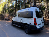 White Winnebago Revel 4x4 RV rental parked on asphalt, surrounded by tall pine trees, ready for adventure.
